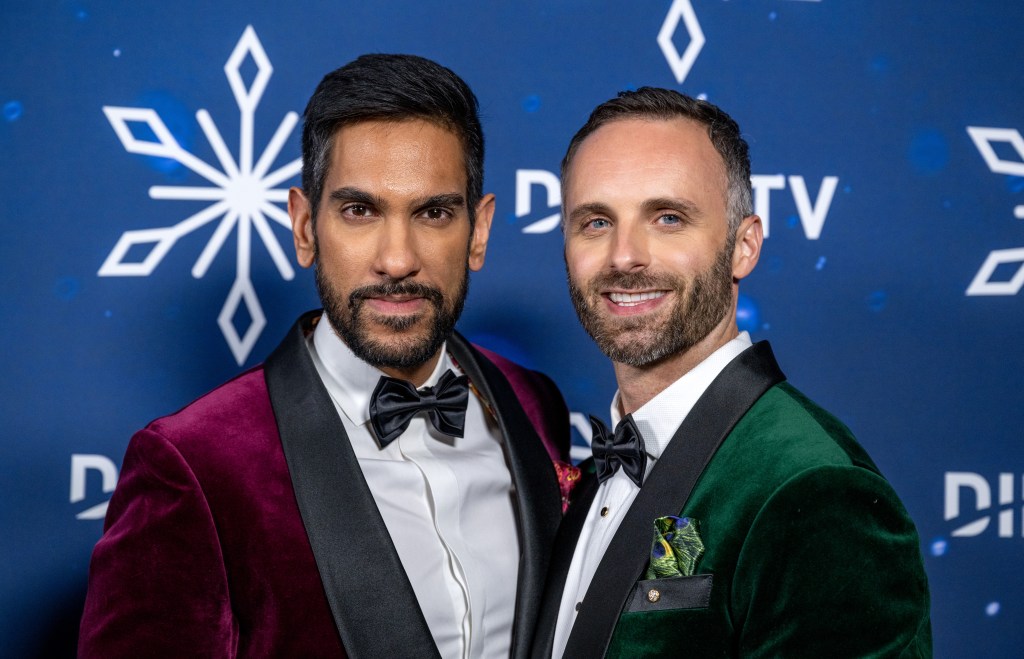 Amrit Kapai in a red blazer standing with husband Nicholas Kouchoukos who's wearing a green blazer