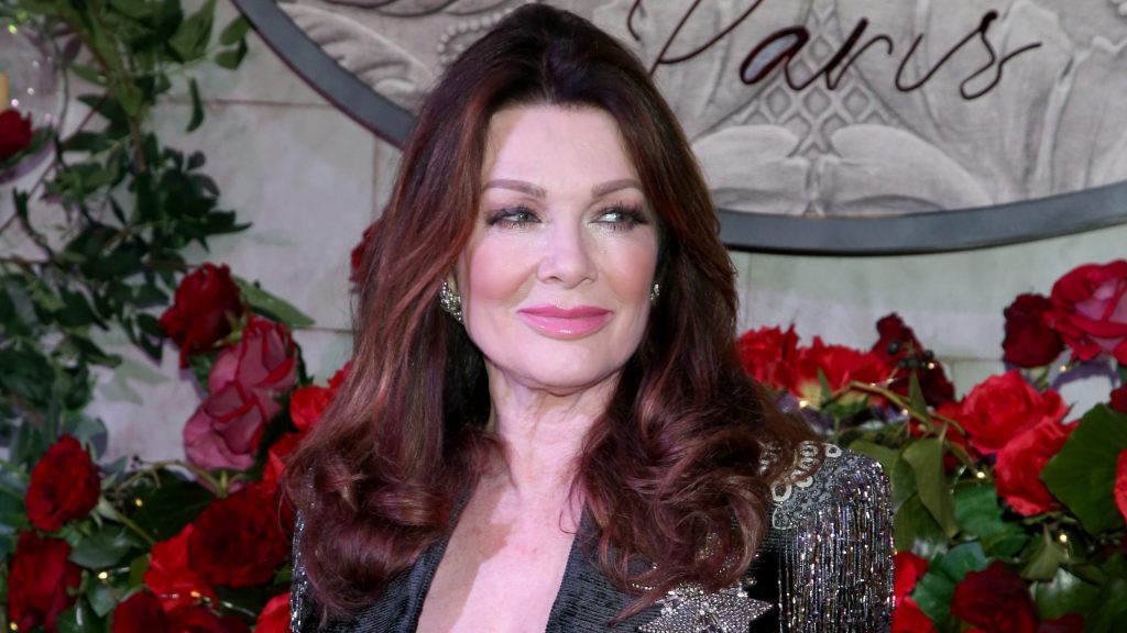 Lisa Vanderpump smiling in a black and silver blazer, standing in front of red flowers