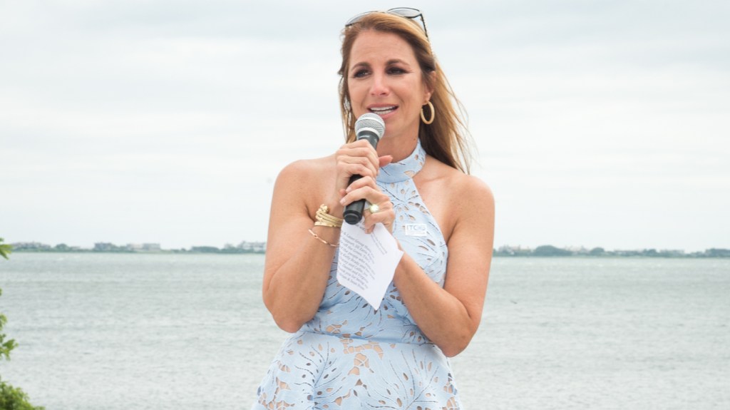 Jill Zarin in a blue dress standing in front of water and speaking into a microphone