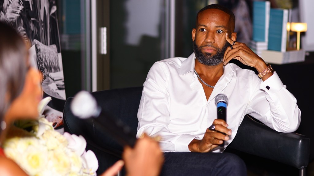 Carlos King in a white button down shirt, resting his head in his hand.