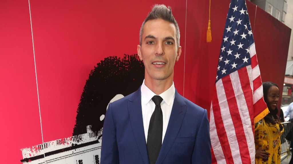 Ari Shapiro in a navy blue suit standing in front of a USA flag