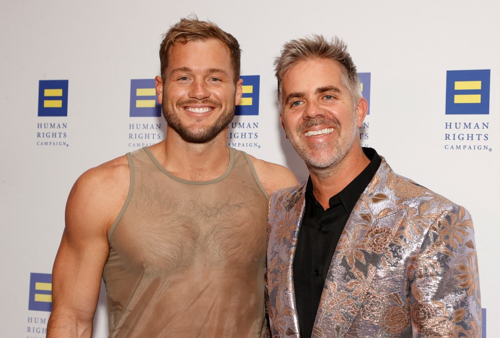 Colton Underwood in a sheer tank top posing with husband Jordan C. Brown who is wearing a multicolored blazer