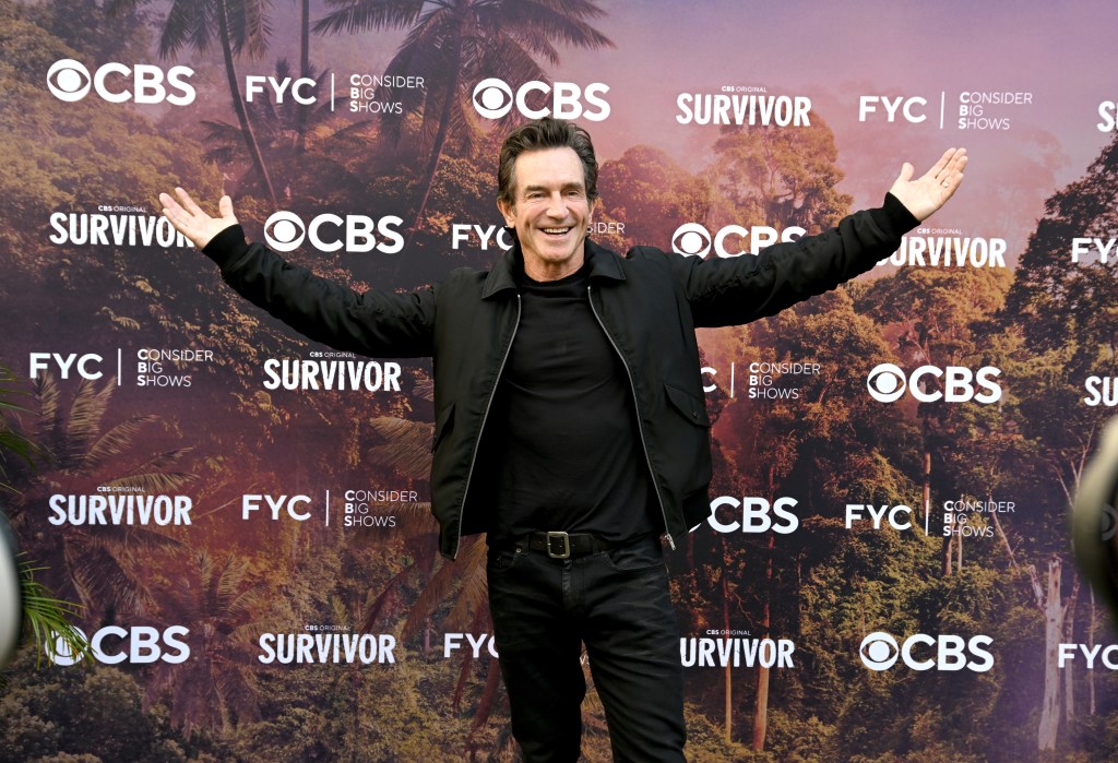 Jeff Probst in an all-black outfit holding his hands up in front of a Survivor backdrop