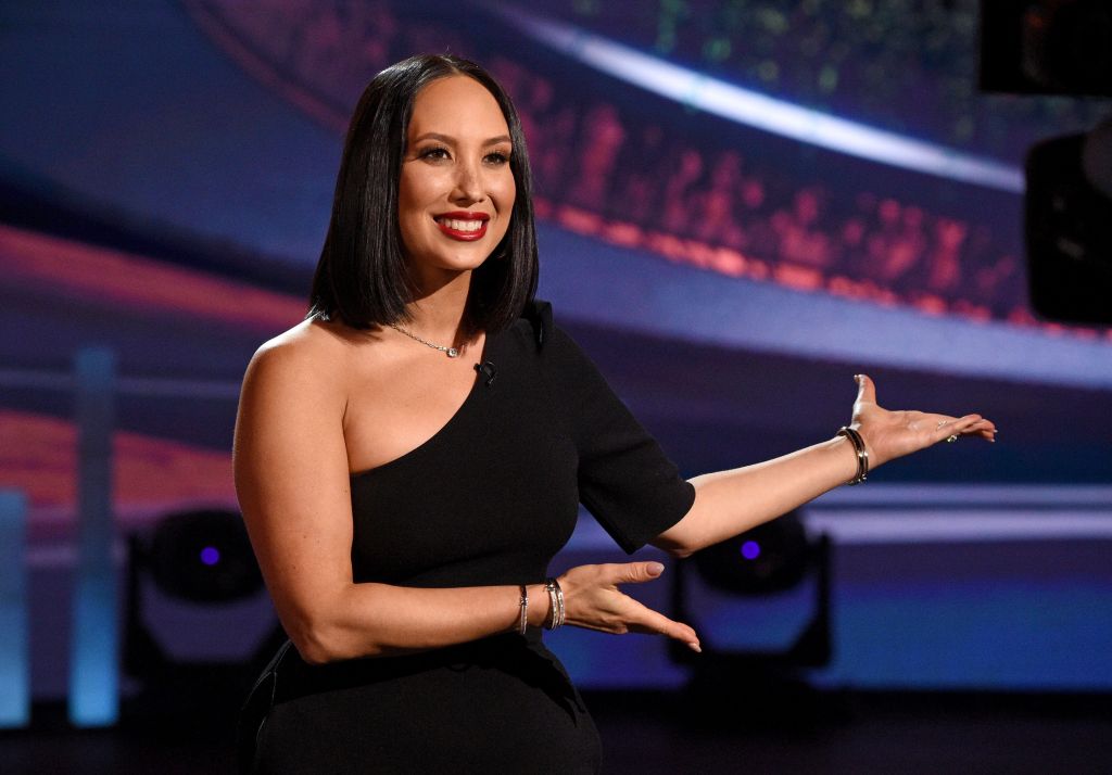 Cheryl Burke sitting in a black dress, holding her arms out to present something