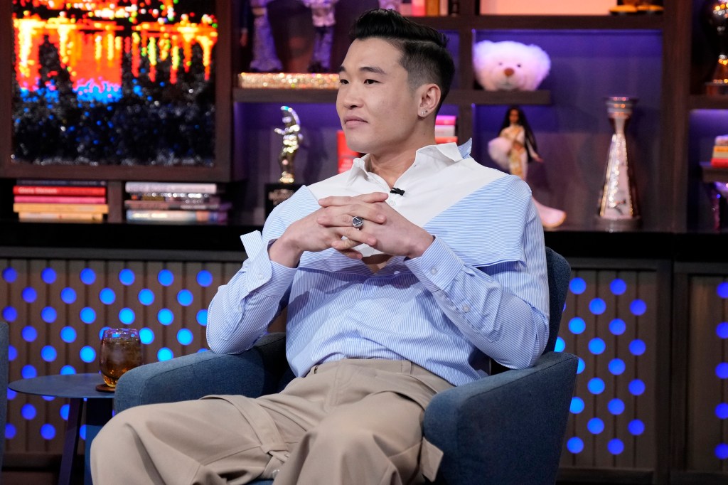 Joel Kim Booster on Watch What Happens Live, wearing a blue shirt sitting with his hands folded