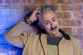 Alan Cumming posing in front of a brick wall while scratching his head